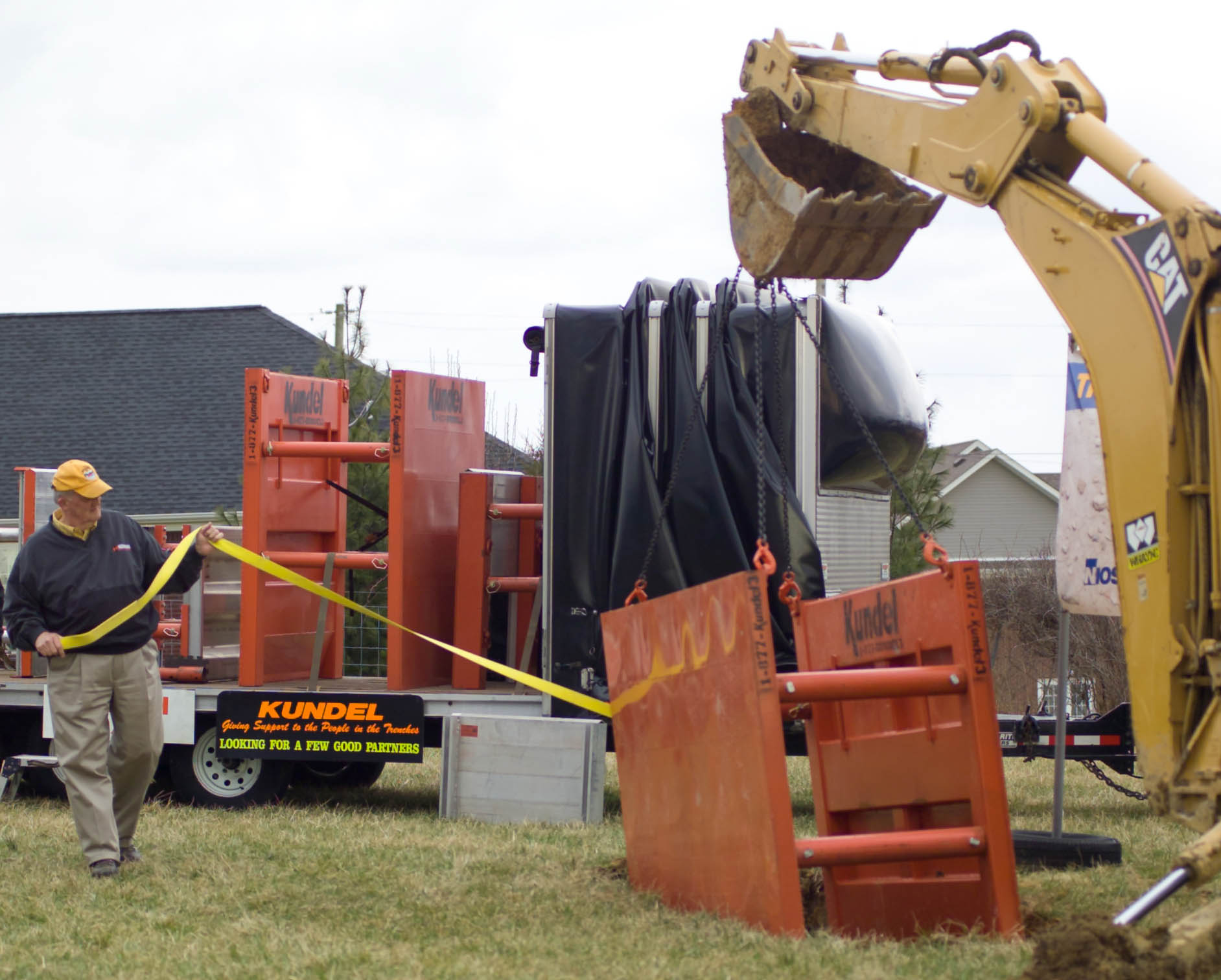 A worker guides a steel trench protection box into an open excavation