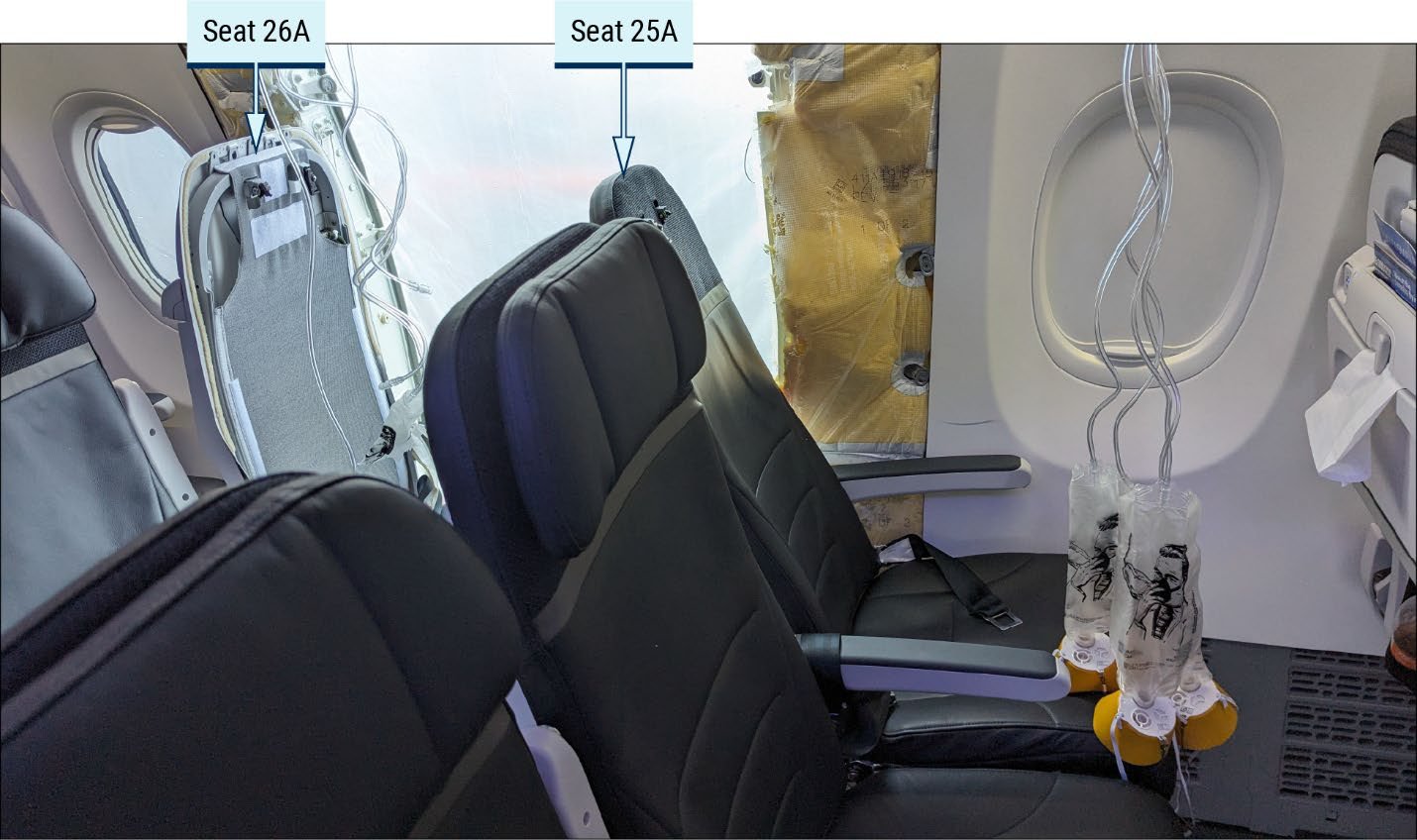 Interior of the airplane cabin after the accident, showing the opening in the left side of the fuselage at row 26 where the door plug had been, with seats 26A and 25A labeled and oxygen masks hanging from the ceiling