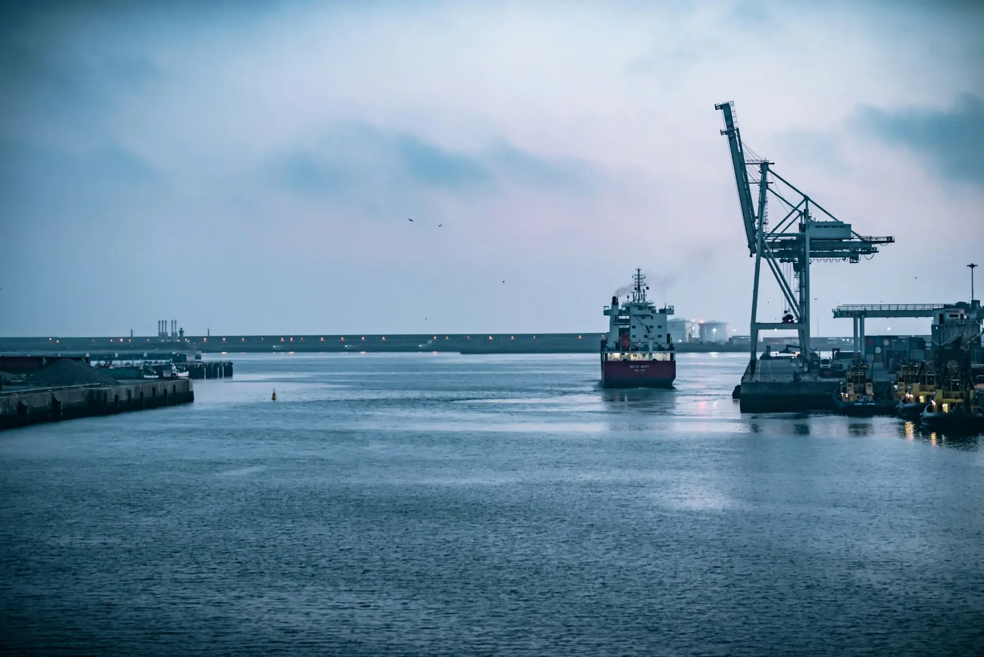 A cargo ship entering a harbor past a container crane at dusk