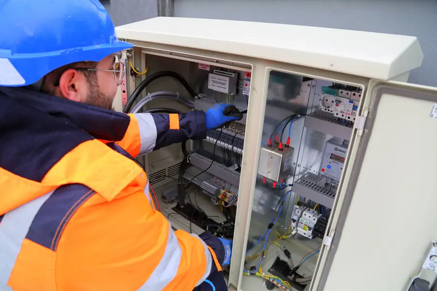 A technician in a hard hat and safety glasses working inside an open electrical control cabinet